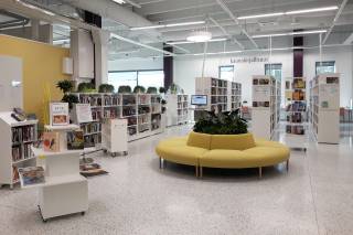 Kaupunginkirjasto Valon aulatila Kuvassa kirjojen esittelyhyllyjä, kirjahyllyjä sekä keltainen, pyöreä sohva, jonka keskellä viherkukkia.