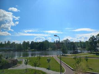 Vehreä puistonäkymä, taustalla kanava. Taivaalla poutapilviä ja vaaleansininen taivas. Aurinko pilkistää pilven takaa.