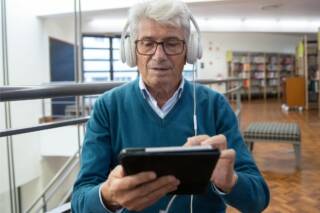 Seniori-ikäinen henkilö selaa tablettia kuulokkeet päässä, taustalla kirjastotilaa.